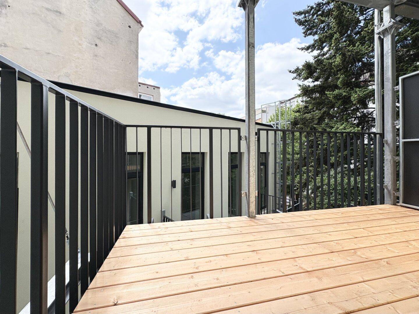 Balkon mit Holzplankenboden und Metallgeländer, umgeben von Bäumen und Gebäuden.