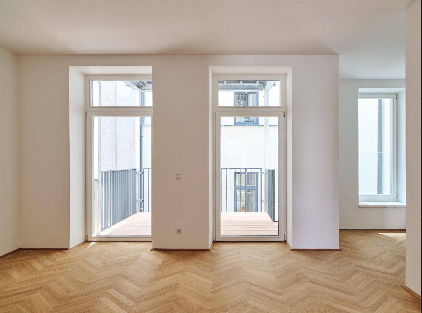 Heller Raum mit Fischgrätparkett und großen Fenstertüren, die auf einen Balkon führen.