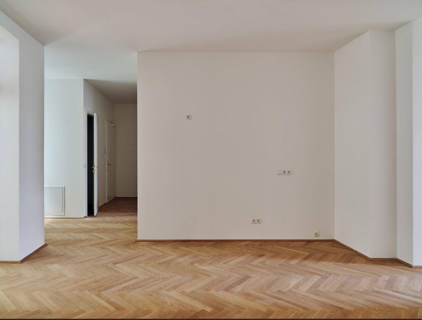 Geräumiges, helles Zimmer mit klassischem Fischgrätparkett und schlichten weißen Wänden.
