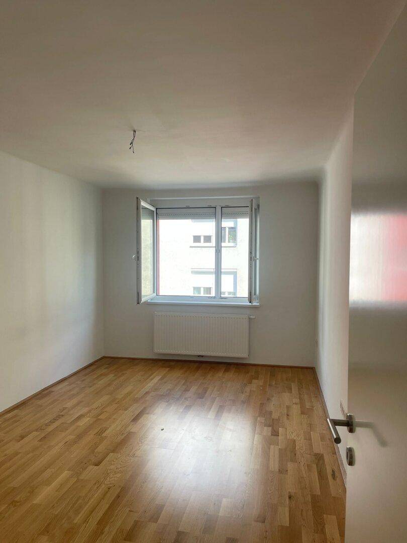 Helles, leeres Zimmer mit Holzparkettboden und einem Fenster mit Blick auf gegenüberliegende Gebäude.