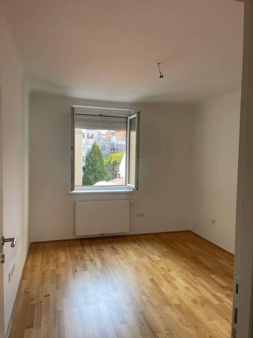Leeres Zimmer mit hellem Parkettboden und Fenster, das einen grünen Ausblick bietet.