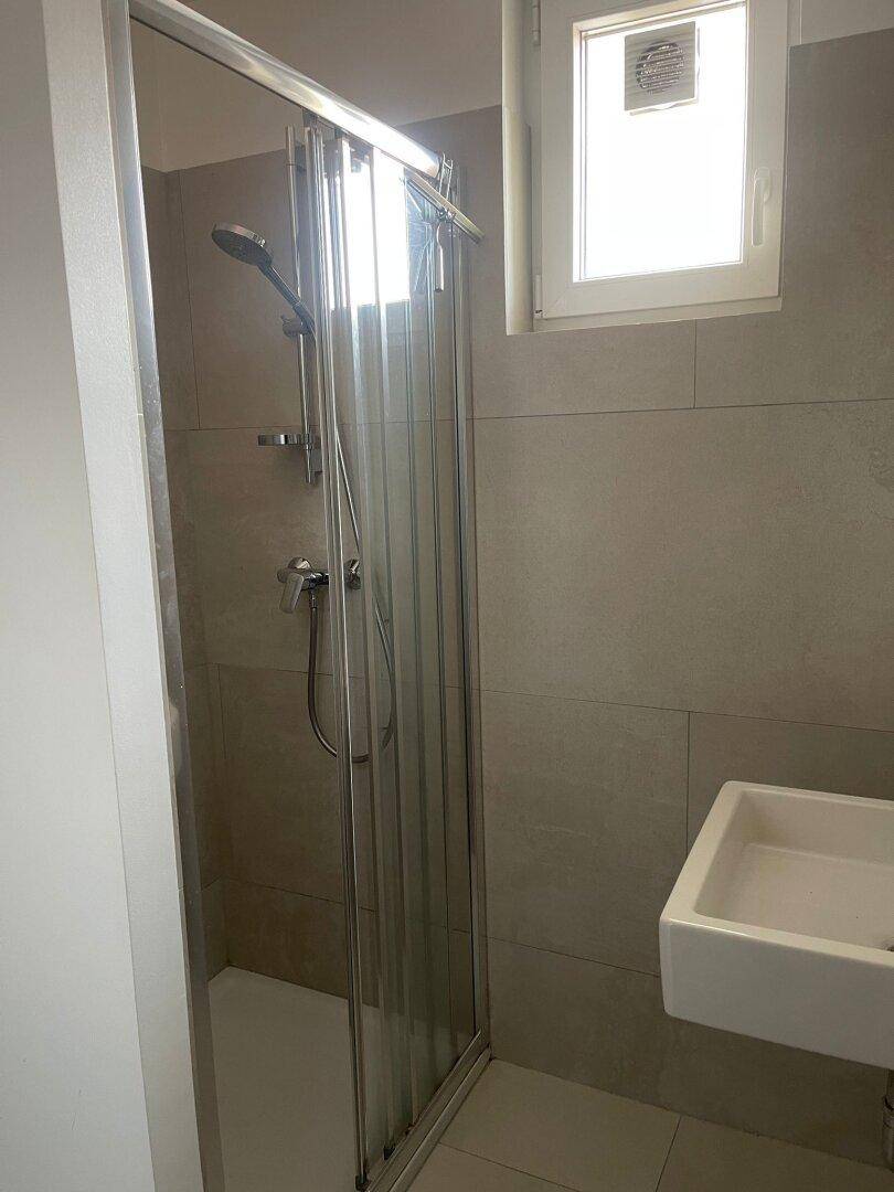 Moderne Duschkabine mit Glasabtrennung und großformatigen Fliesen an den Wänden, sowie einem Fenster.