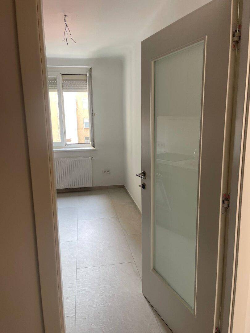 Blick durch eine Tür in ein helles Zimmer mit Fliesenboden und Fenster.