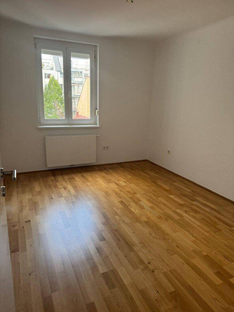 Helles, leeres Zimmer mit Holzparkettboden und Fenster mit Blick ins Grüne.