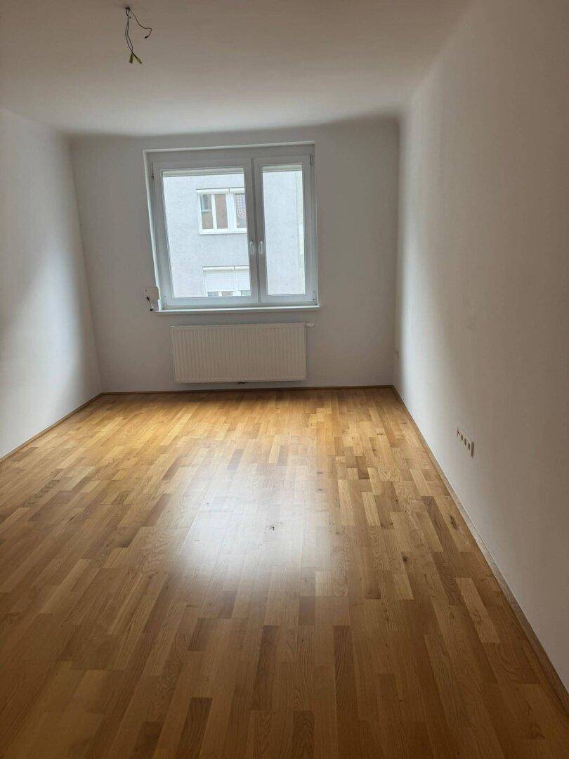 Helles, leeres Zimmer mit Holzparkettboden und Fenster mit Blick auf Gebäude.