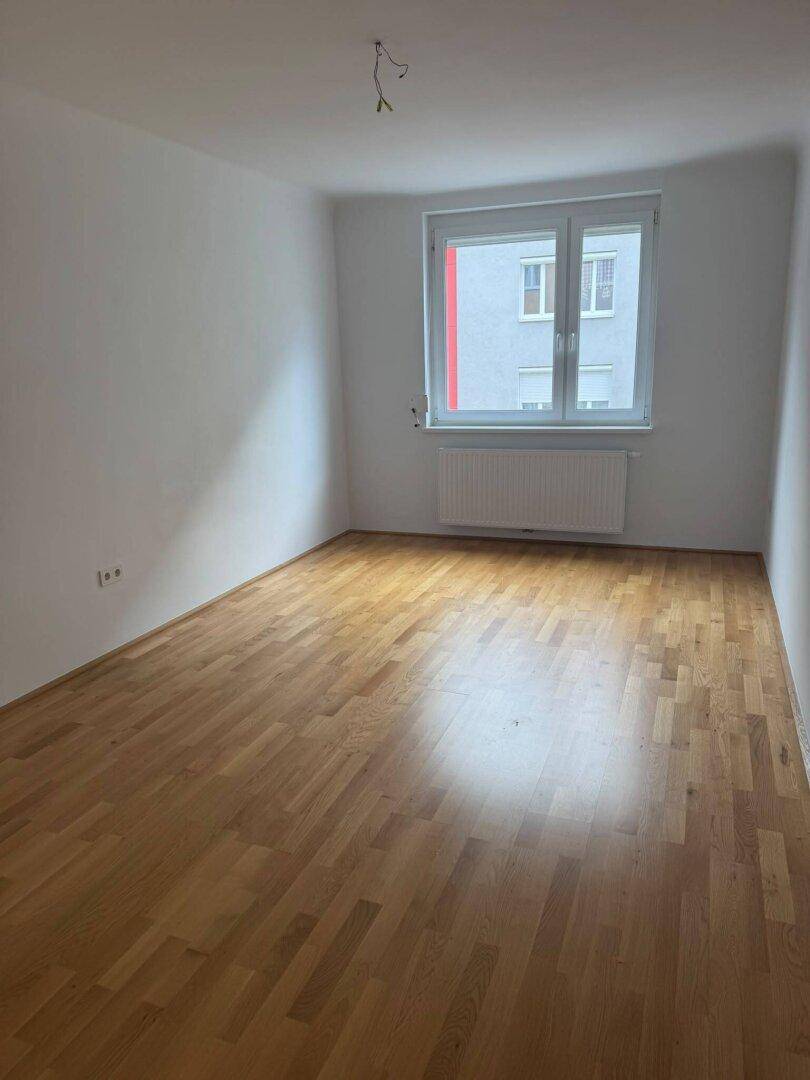 Leeres Zimmer mit hellem Parkettboden und Fenster, das viel Tageslicht hereinlässt.