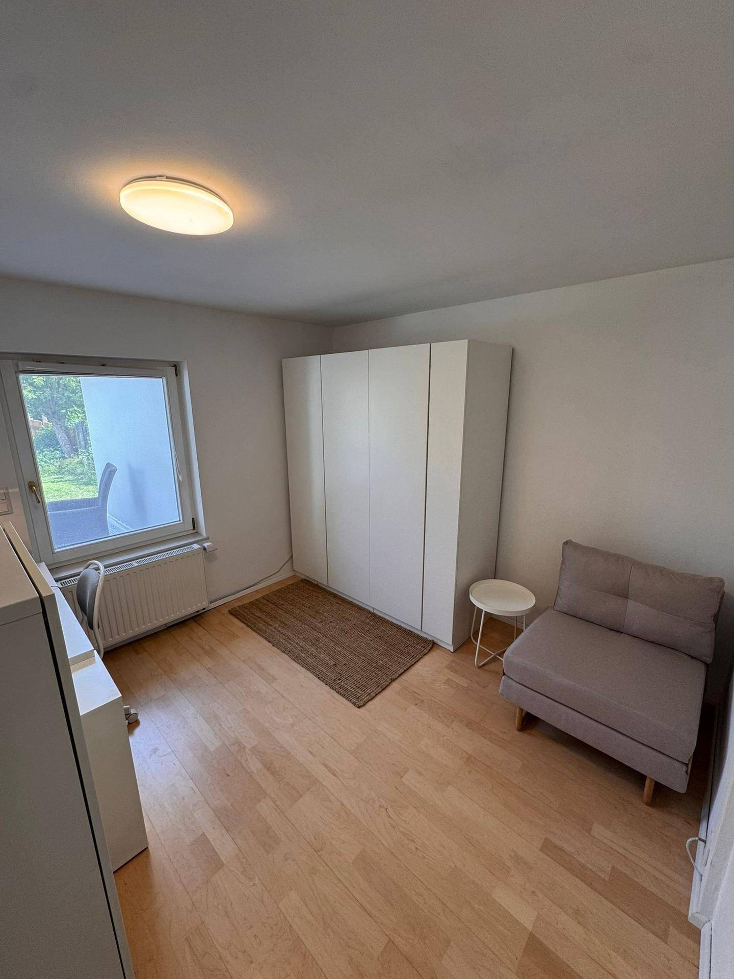 Helles Zimmer mit Fenster, Kleiderschrank und einer kleinen Sitzgelegenheit.