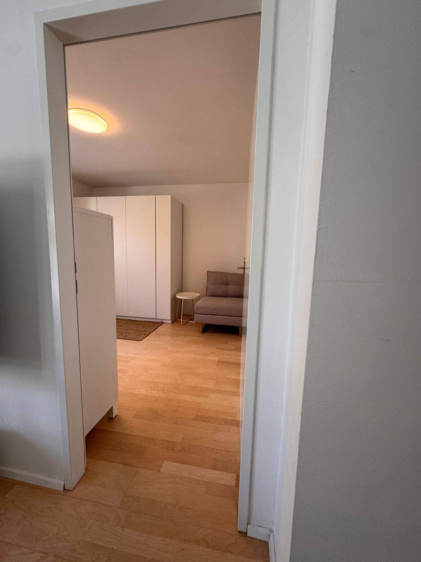 Blick durch eine Tür in einen hellen Raum mit Schrank und Sitzgelegenheit.