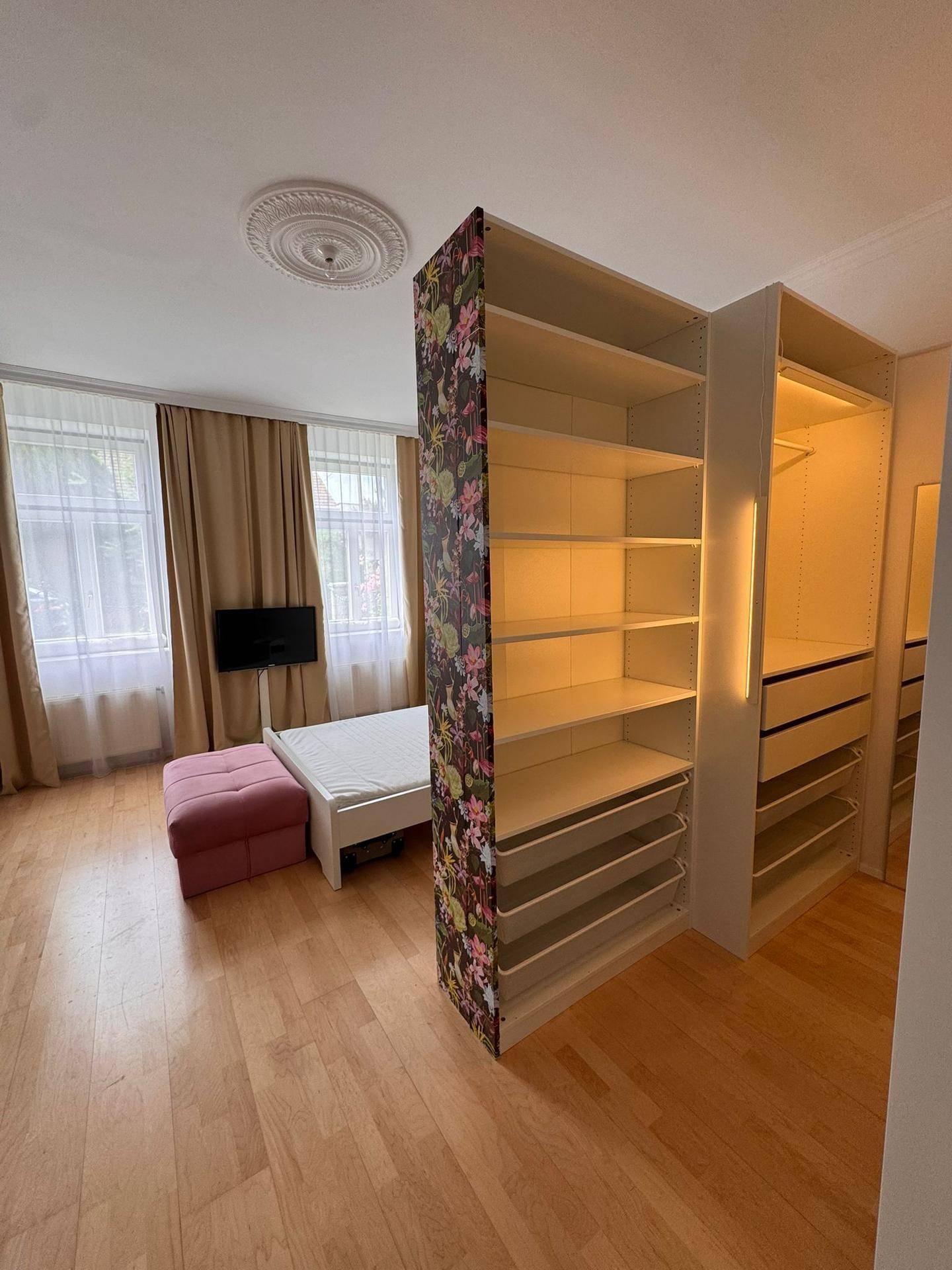 Schlafzimmer mit Fenster, Fernseher und offenem Regalsystem hinter dem floralen Raumteiler.