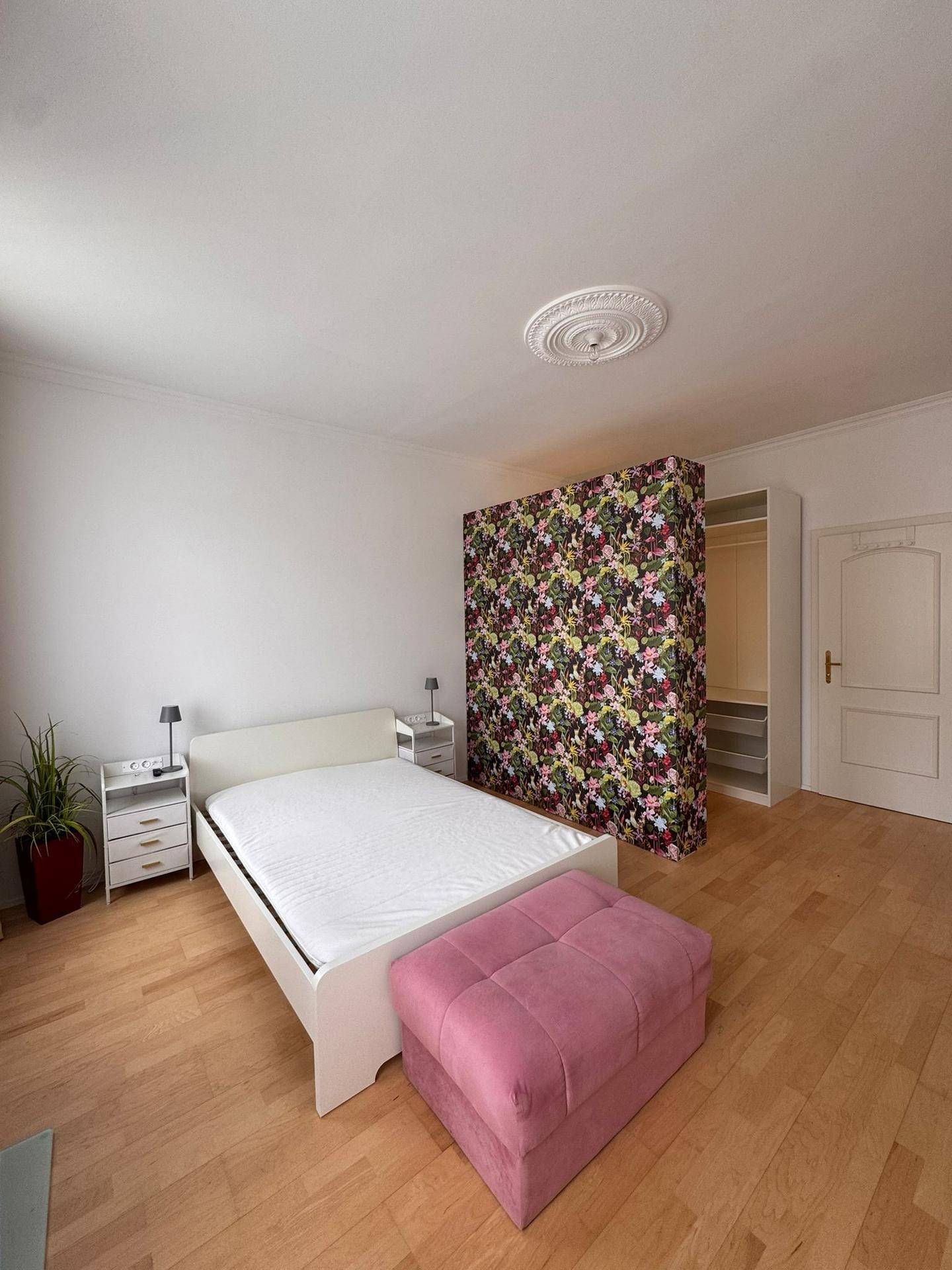Geräumiges Schlafzimmer mit Doppelbett, rosa Ottomane und floral gemustertem Raumteiler.