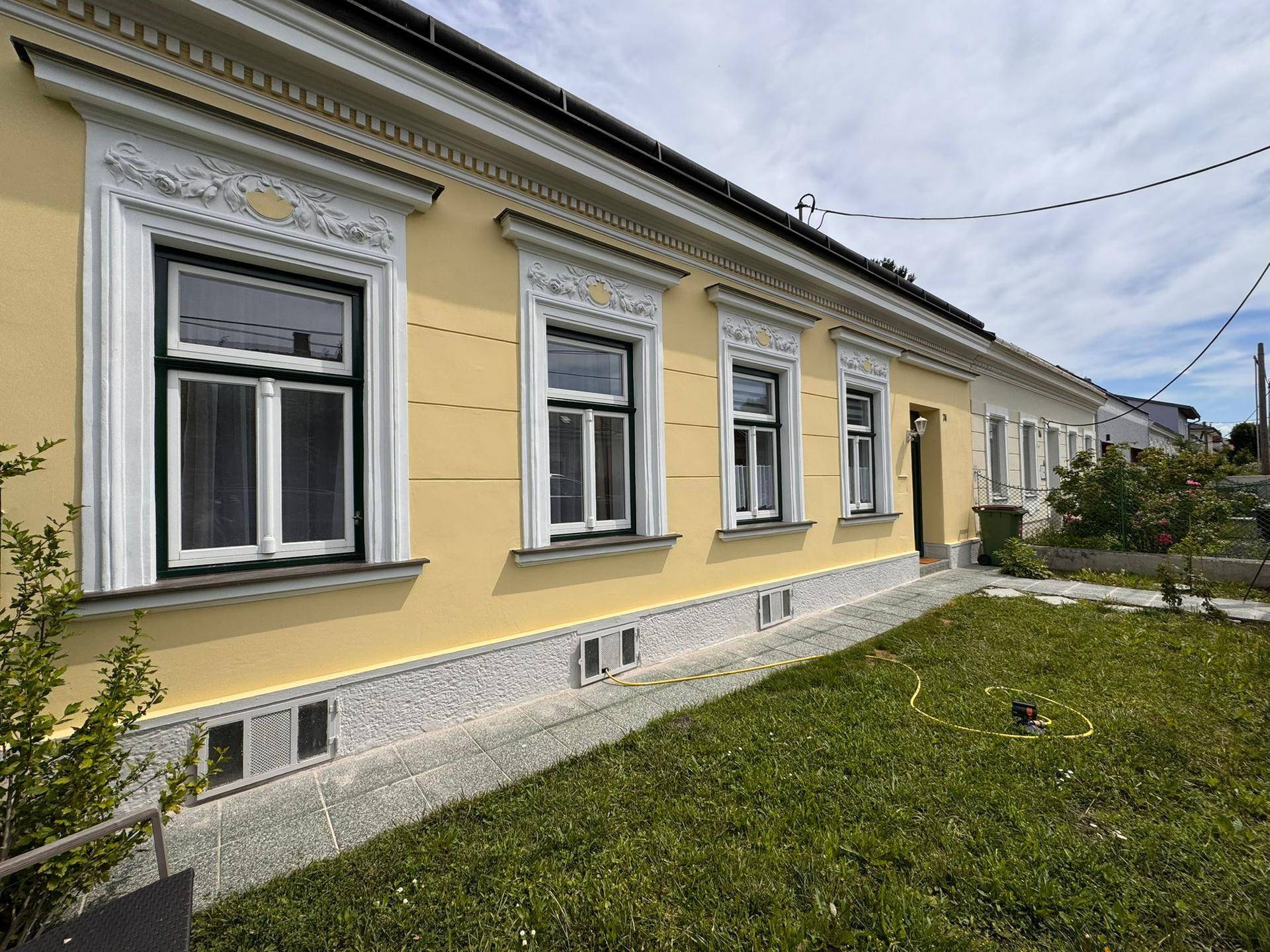 Seitenansicht des Hauses mit gelber Fassade, weißen Fenstern und kleinem Rasenbereich.