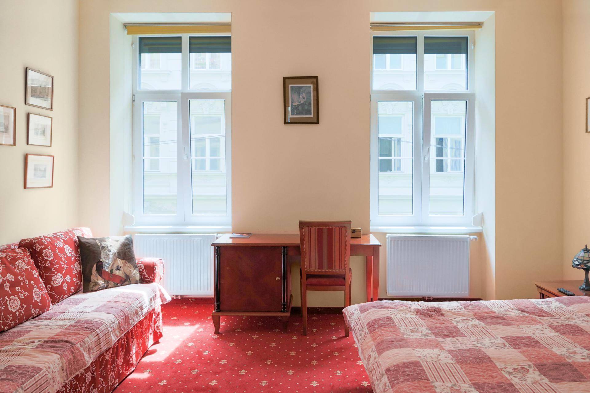 Gemütliches Schlafzimmer mit rotem Teppichboden, zwei Fenstern und einem bequemen Sofa.