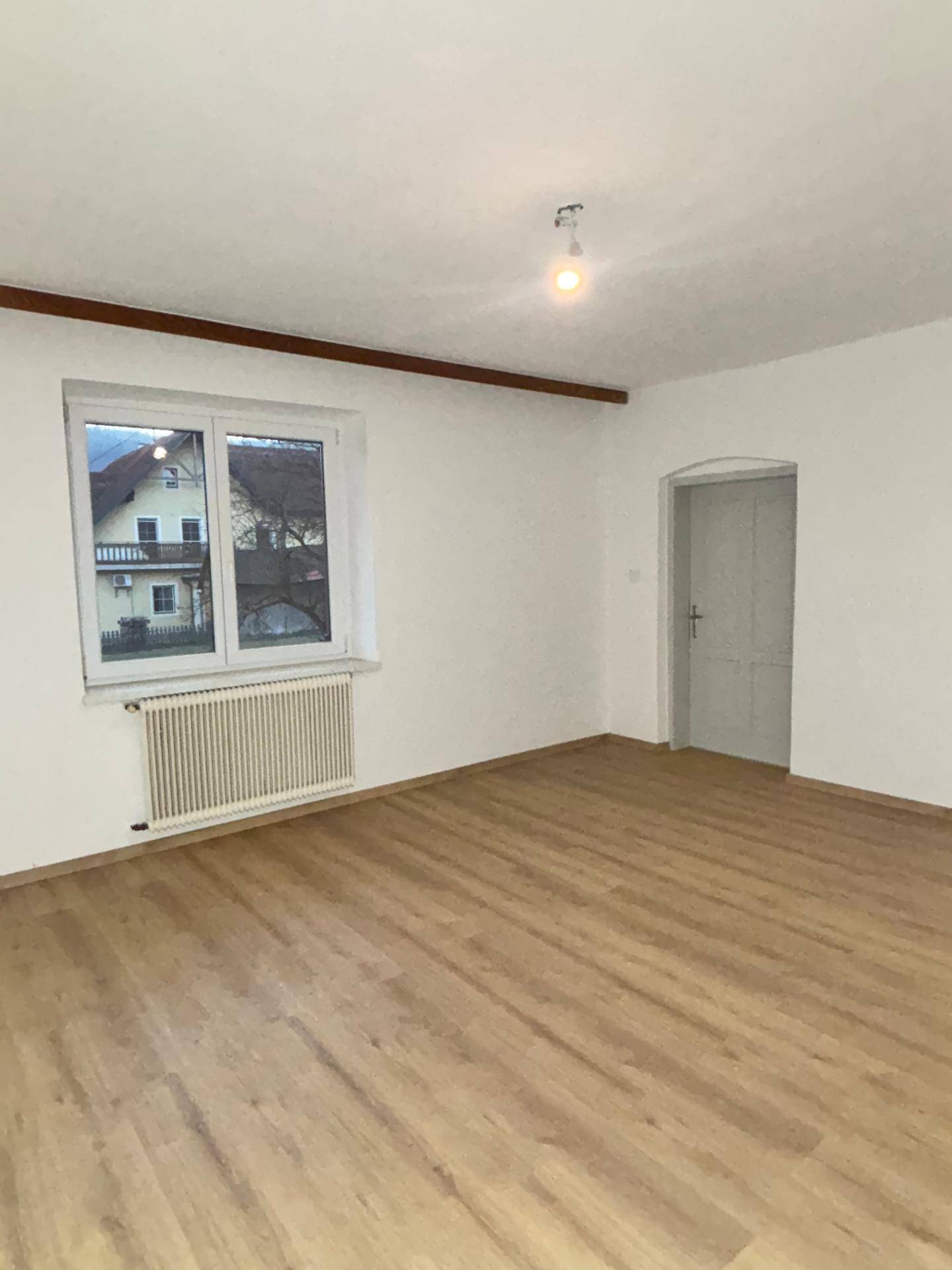 Leerer Raum mit Laminatboden, Fenster, Heizkörper und Holzbalken an der Decke.