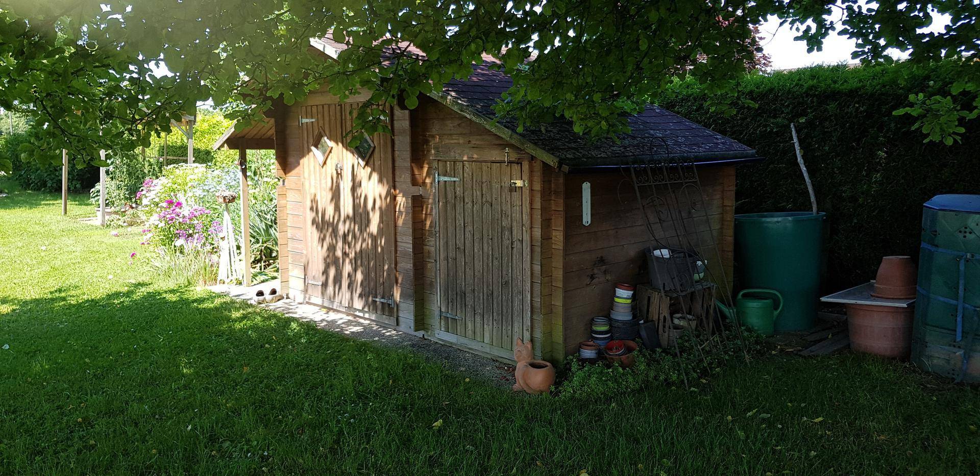 Kleines Holzgartenhaus mit Satteldach, umgeben von Rasen und Gartenutensilien, unter einem Baum.