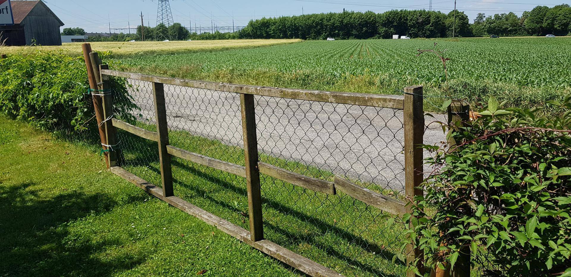 Ländliche Szenerie mit einem Holzzaun, der eine Wiese von einem Feld trennt, unter blauem Himmel.