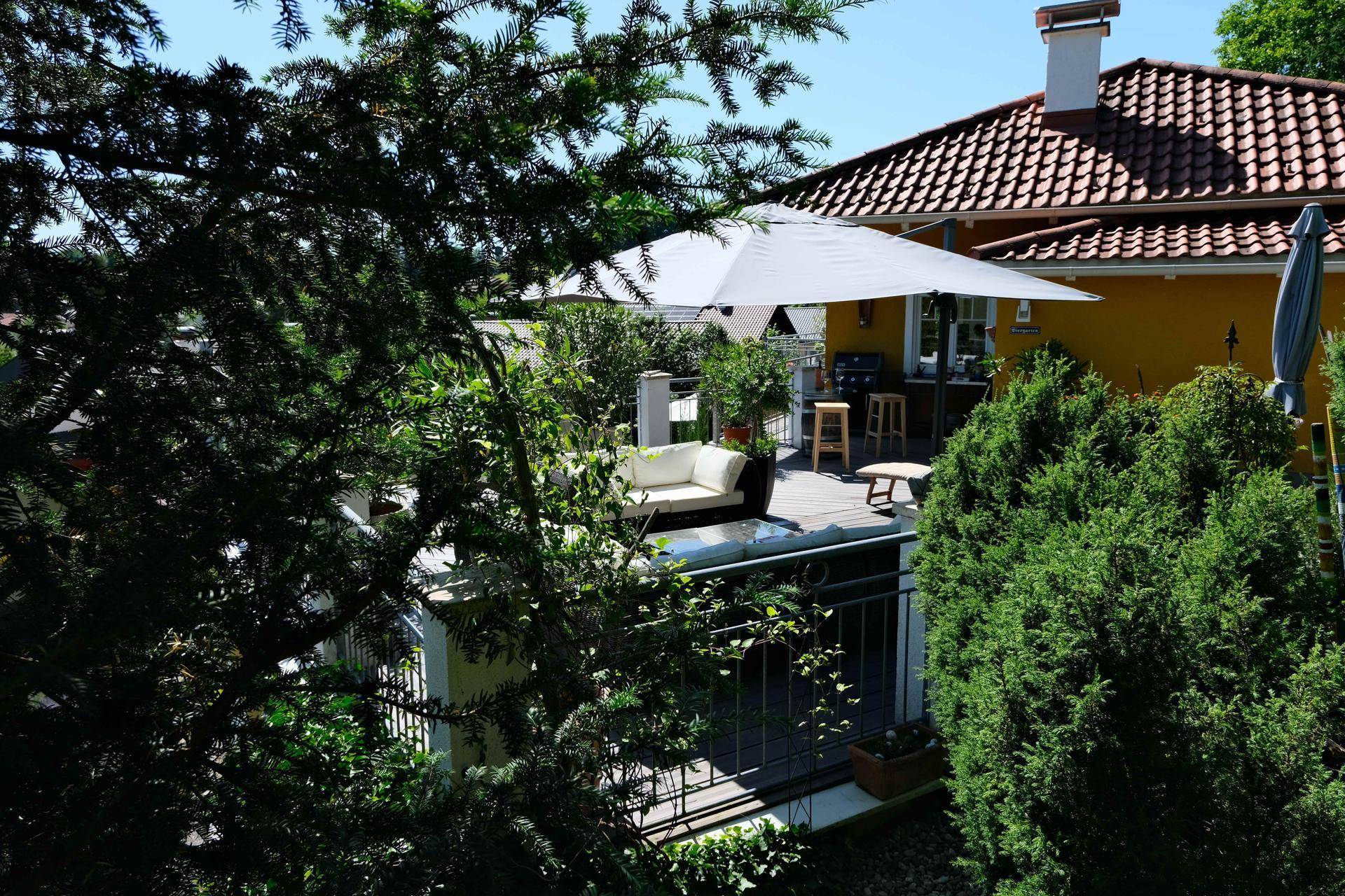 Blick durch Bäume auf eine sonnige Terrasse mit Sitzgelegenheiten und Sonnenschirm.