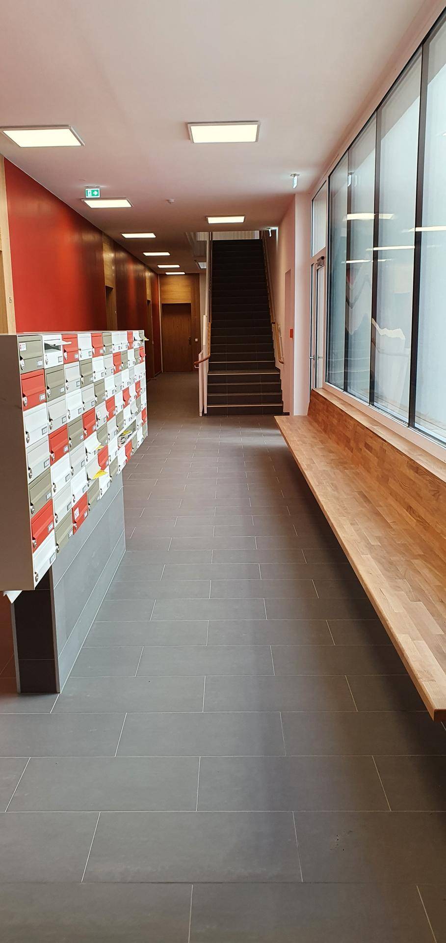 Heller Gemeinschaftsflur mit großen Fenstern, Sitzbank und Schließfächern, führt zu einer Treppe.