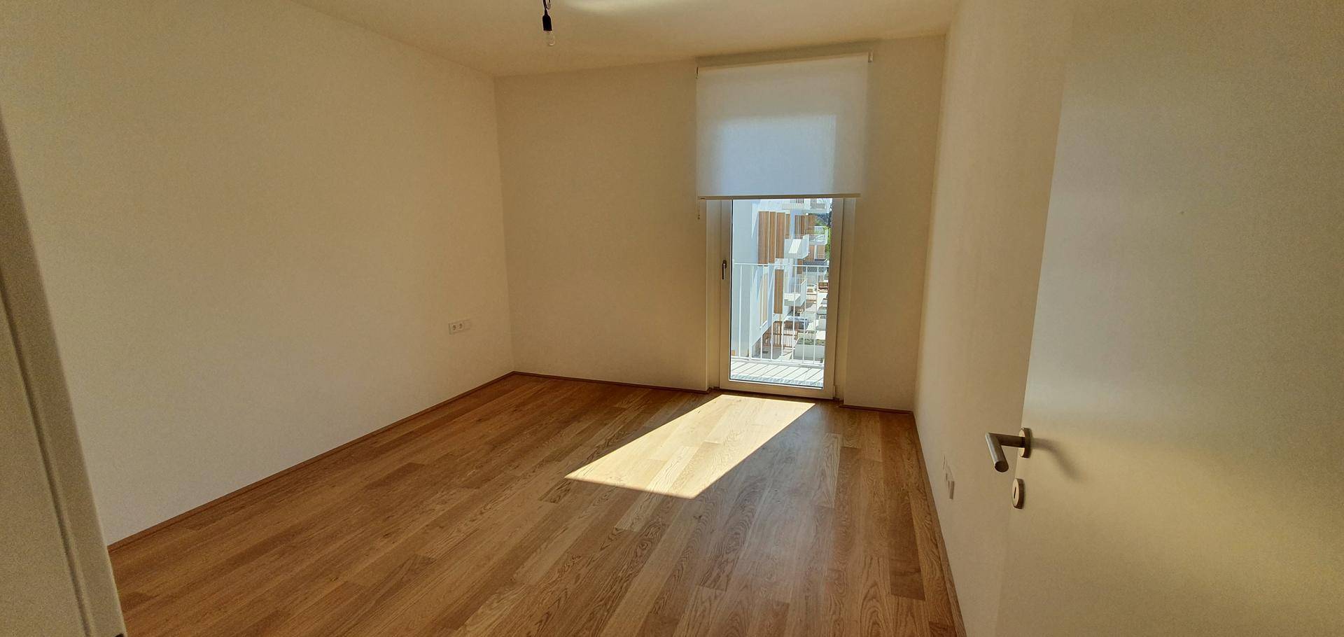 Schlafzimmer mit hellem Holzparkett und Zugang zu einem Balkon, gut beleuchtet durch eine Fenstertür.