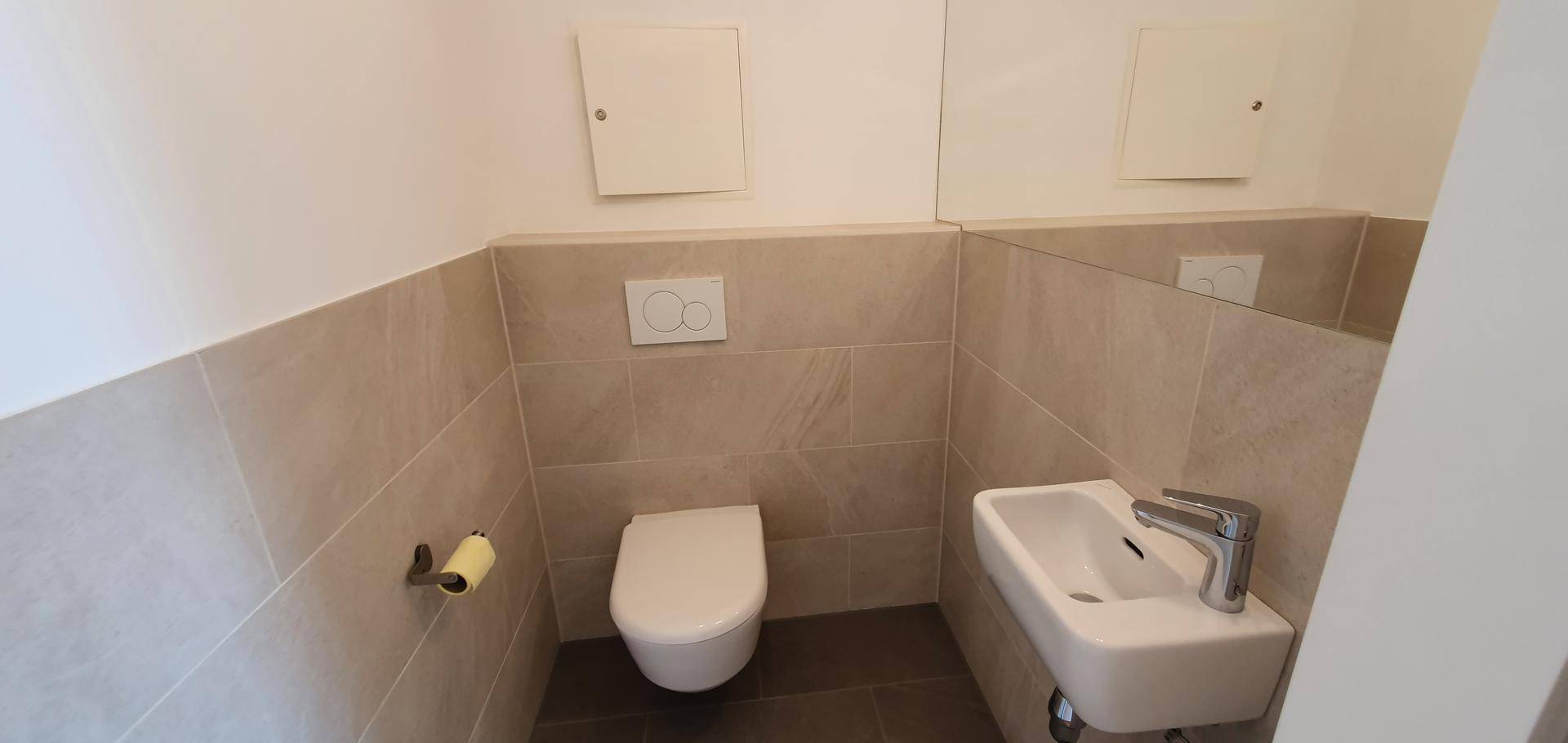 Modernes Gäste-WC mit Wand-WC, kleinem Waschbecken und hellen Fliesen an den Wänden und auf dem Boden.