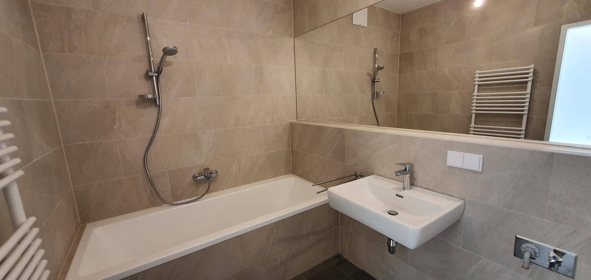 Modernes Badezimmer mit Badewanne, Duscharmatur, Waschbecken und großem Spiegel, ausgestattet mit hellen Fliesen.