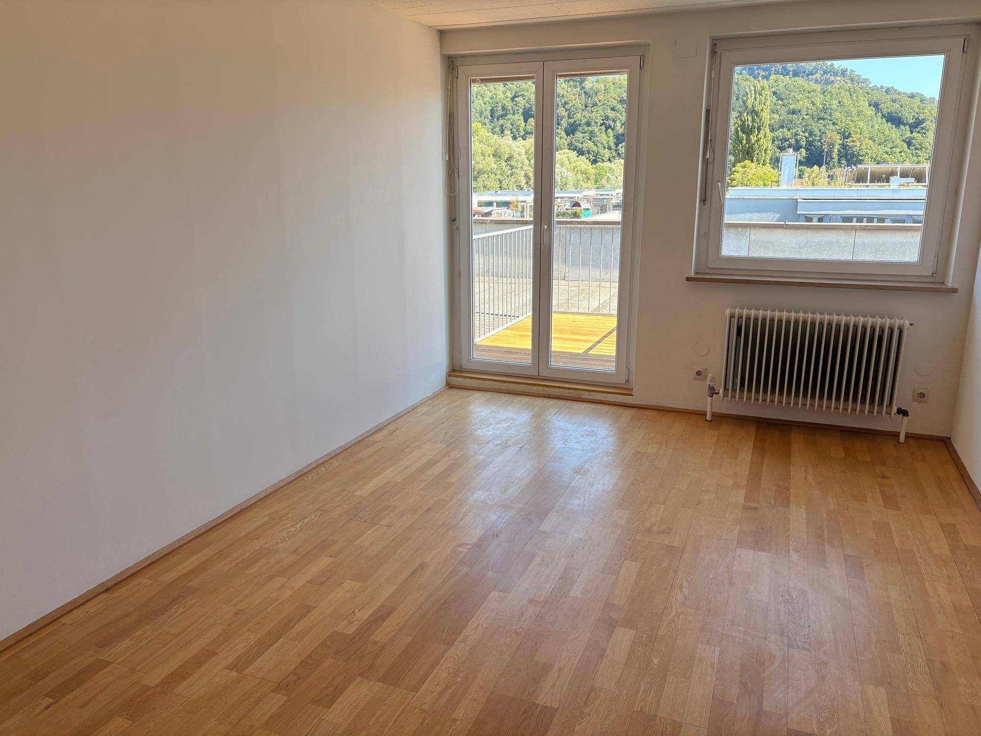 Heller Raum mit Holzboden, weißen Wänden, einer Balkontür und einem Fenster mit Ausblick.
