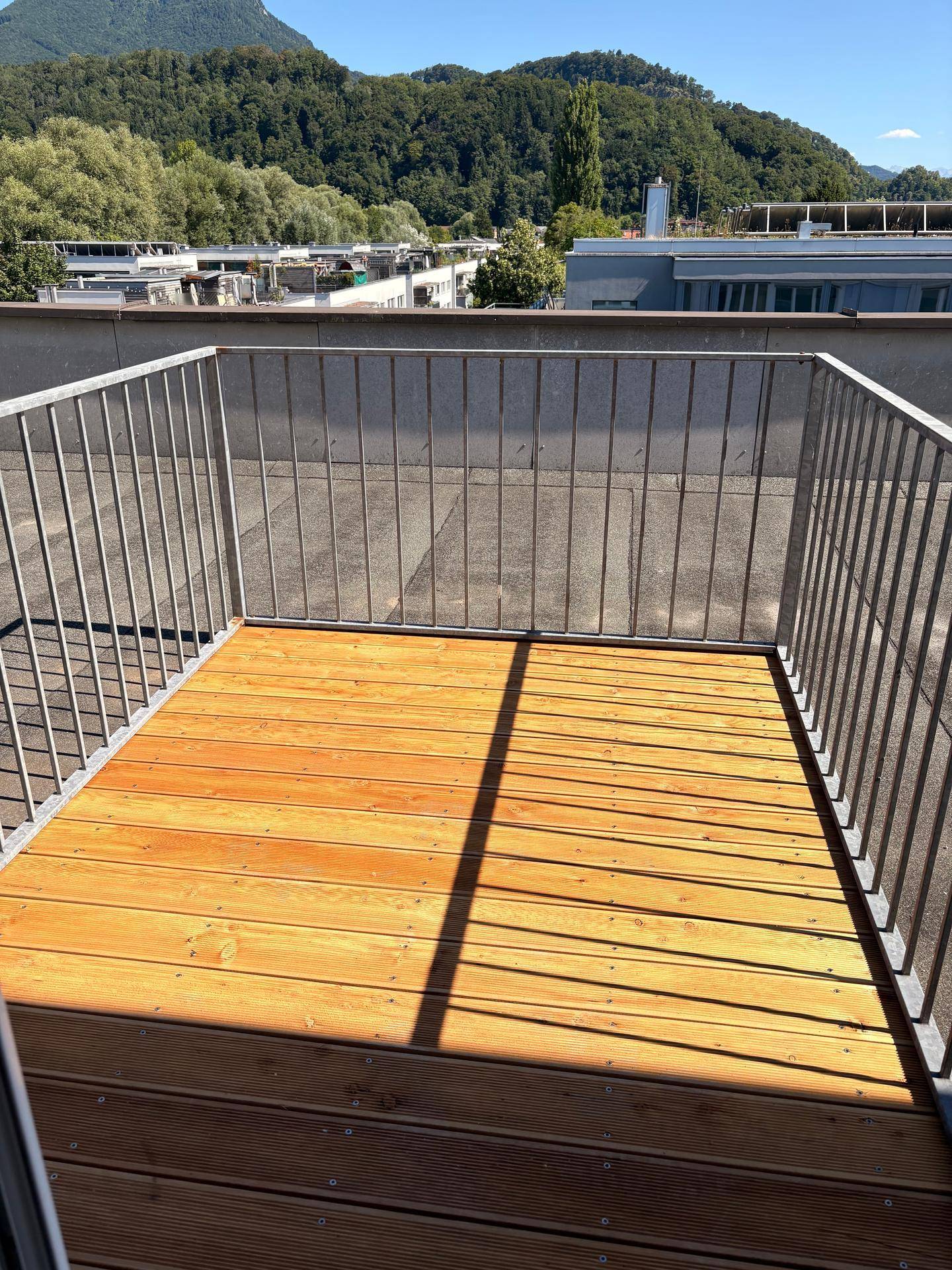 Heller Balkon mit Holzfußboden und Metallgeländer, bietet einen Ausblick auf Dächer und grüne Hügel.