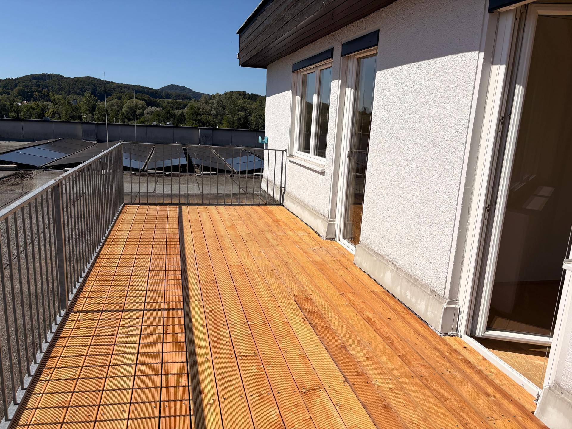Langer Balkon mit Holzfußboden und Metallgeländer, bietet einen weiten Blick über Dächer und grüne Landschaft.