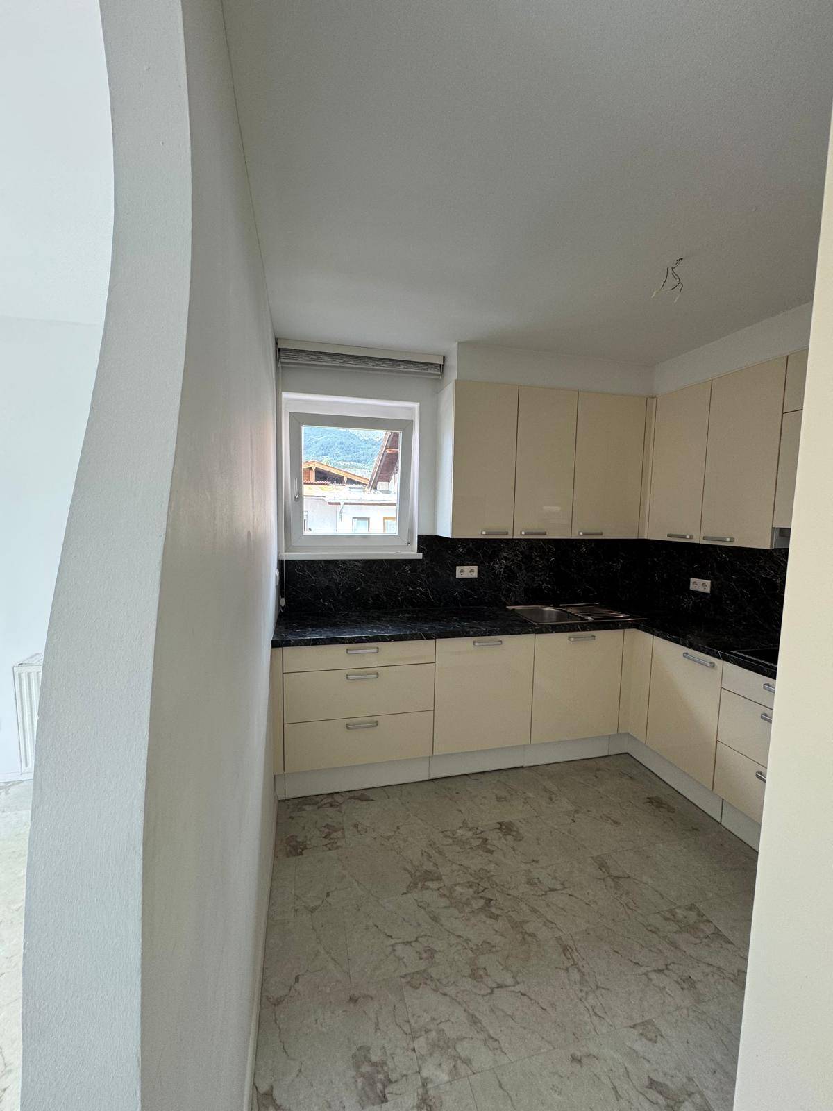 Moderne Einbauküche mit beigen Hochglanzfronten, dunkler Arbeitsplatte und Fenster.
