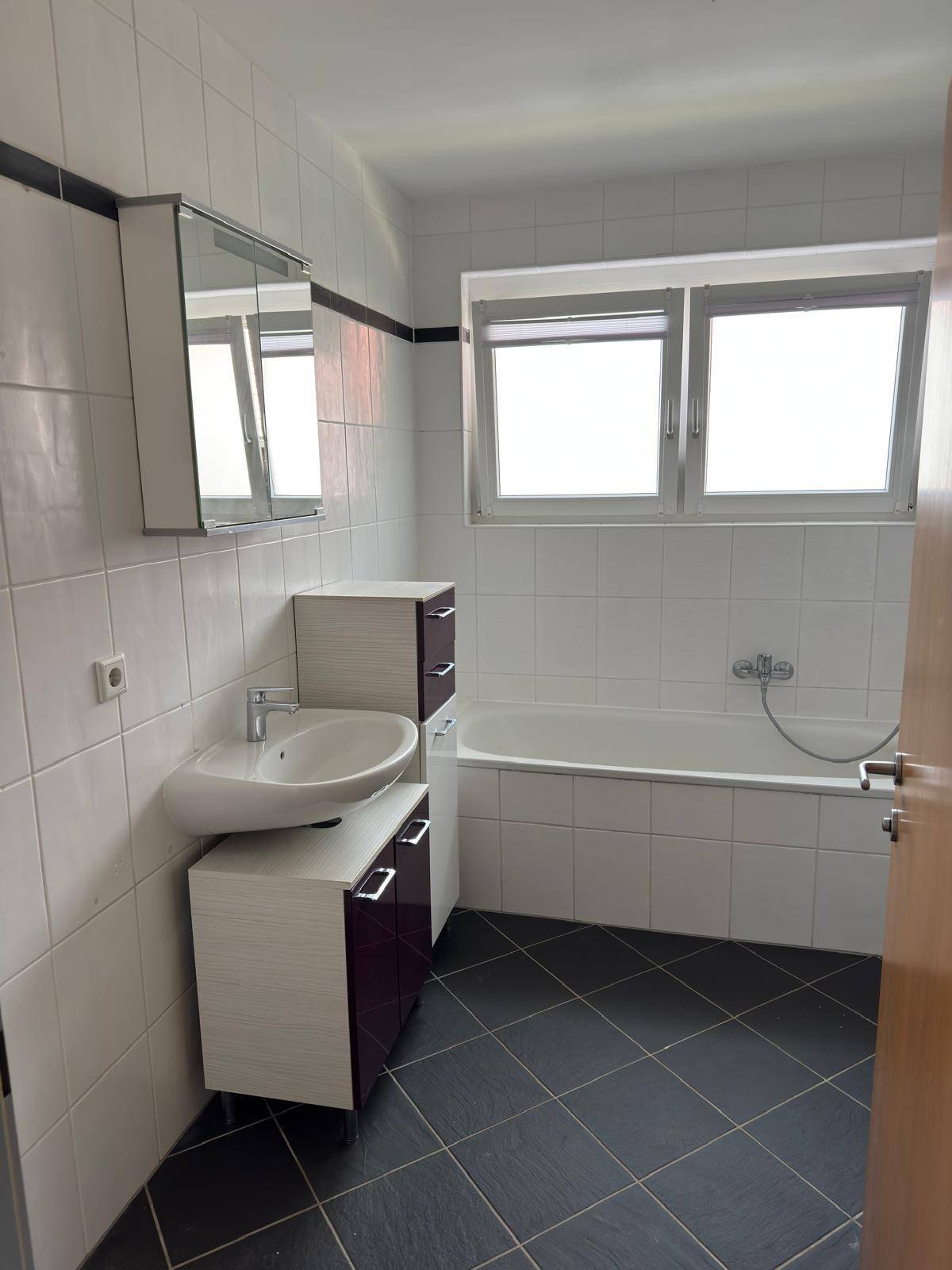 Badezimmer mit Badewanne, Waschbecken, Spiegelschrank und Fenster für Tageslicht.