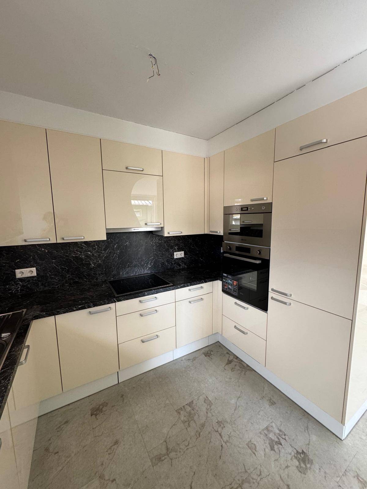 Moderne Einbauküche mit beigen Hochglanzfronten, dunkler Arbeitsplatte und integrierten Geräten.