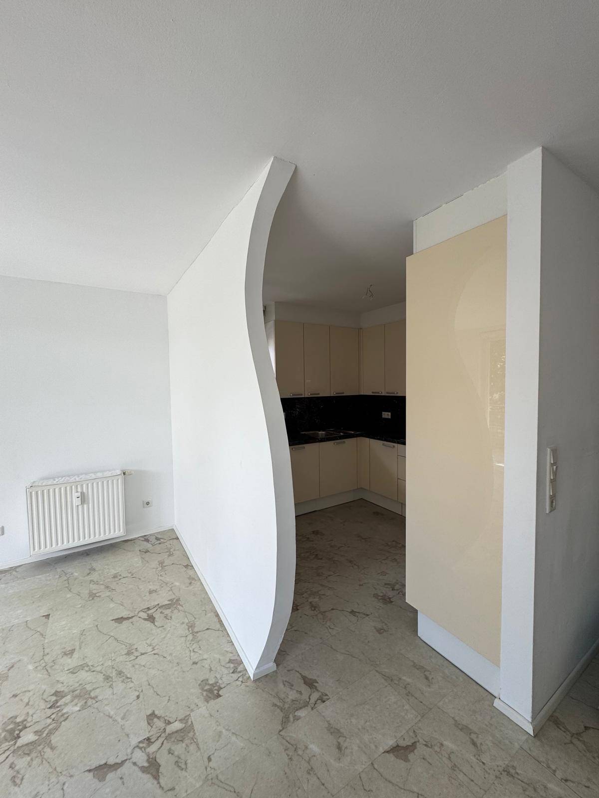 Heller Wohnbereich mit Steinboden und einer geschwungenen Wand, die zur Küche führt.