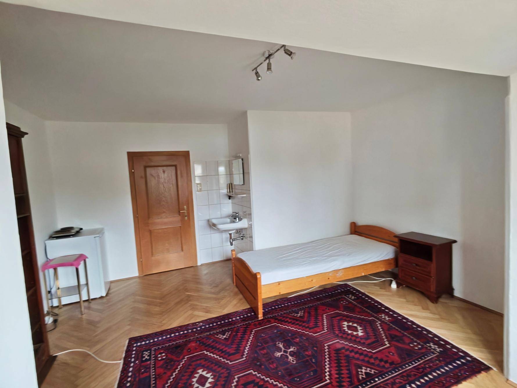 Schlafzimmer mit Parkettboden, Einzelbett, Kühlschrank und integriertem Waschbecken.
