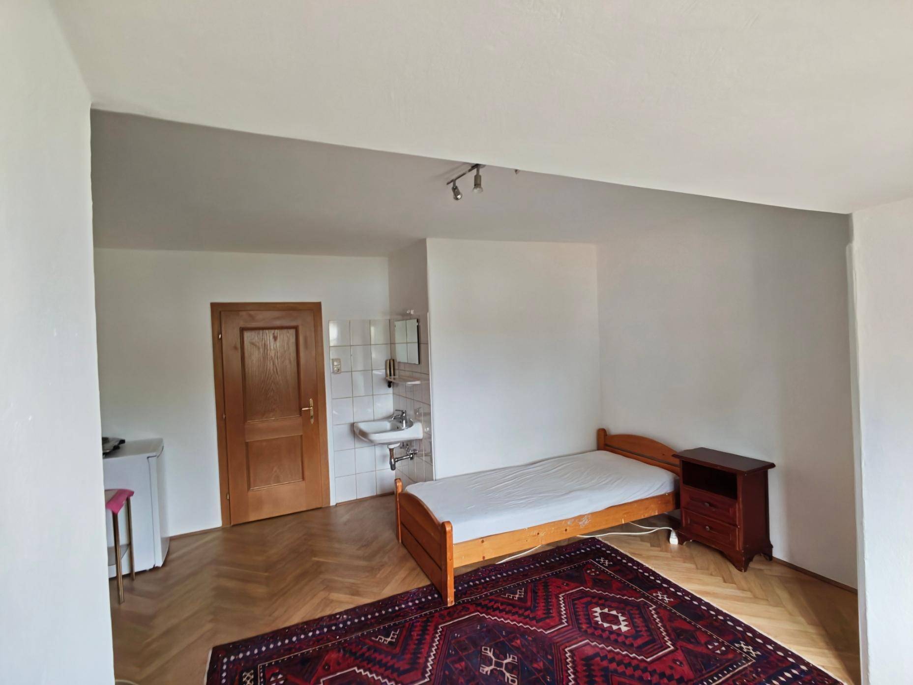Einfaches Schlafzimmer mit Einzelbett, Parkettboden und integriertem Waschbecken.