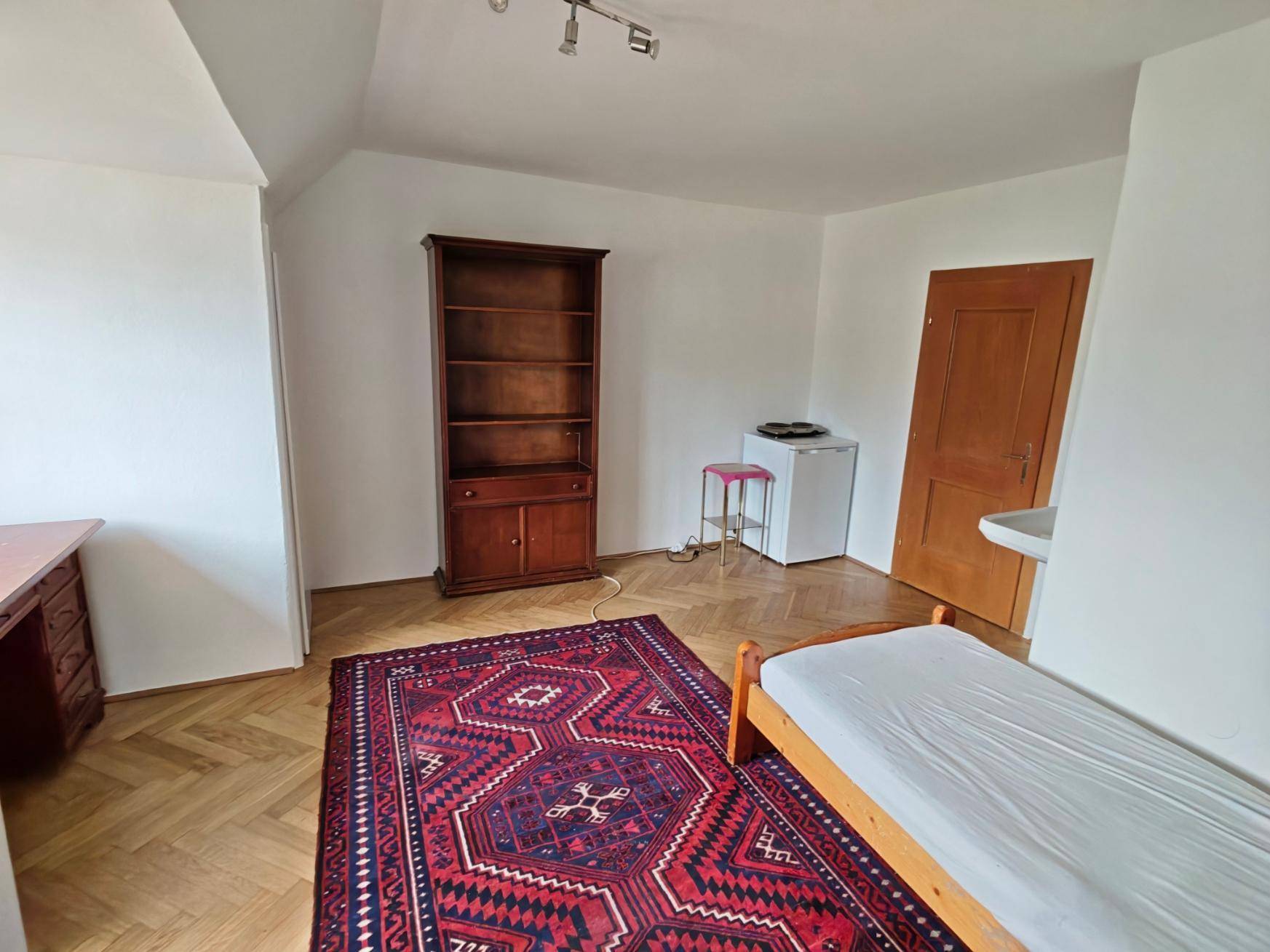 Schlafzimmer mit Parkettboden, Einzelbett, Bücherregal und kleinem Kühlschrank im Raum.