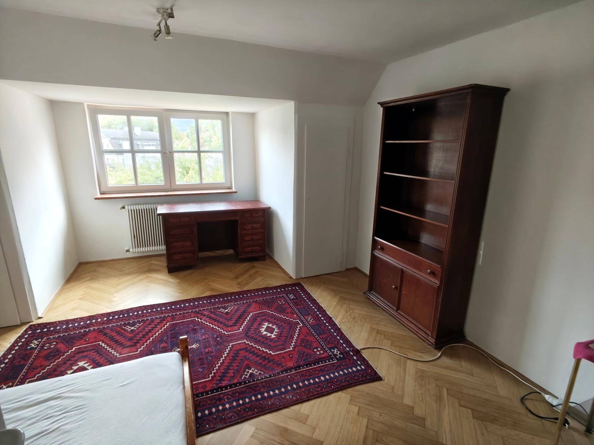 Helles Schlafzimmer mit Parkettboden, Schreibtisch, Bücherregal und Heizkörper unter dem Fenster.