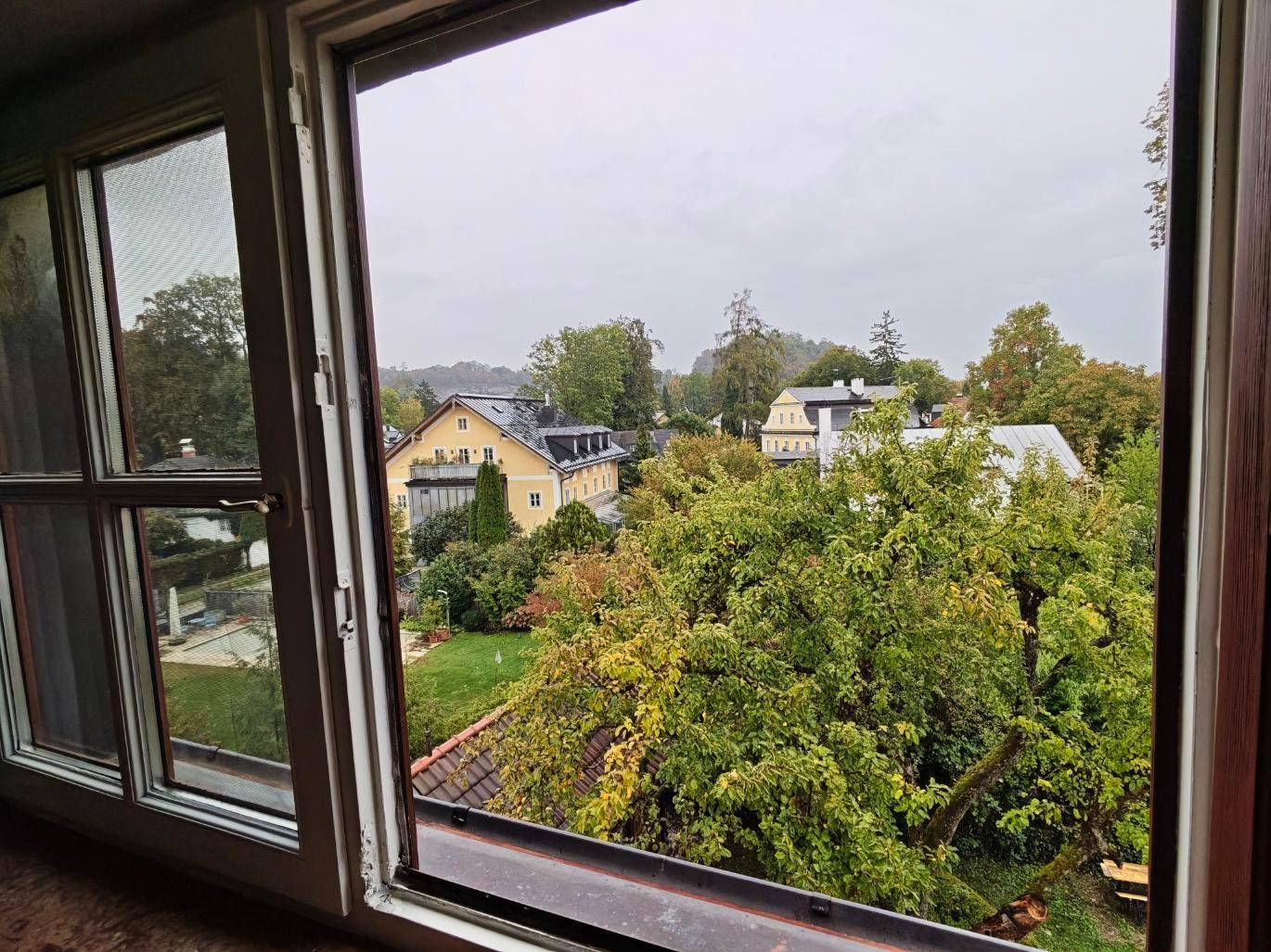 Offener Fensterblick aus dem Schlafzimmer auf eine grüne Umgebung mit weiteren Gebäuden und Natur.