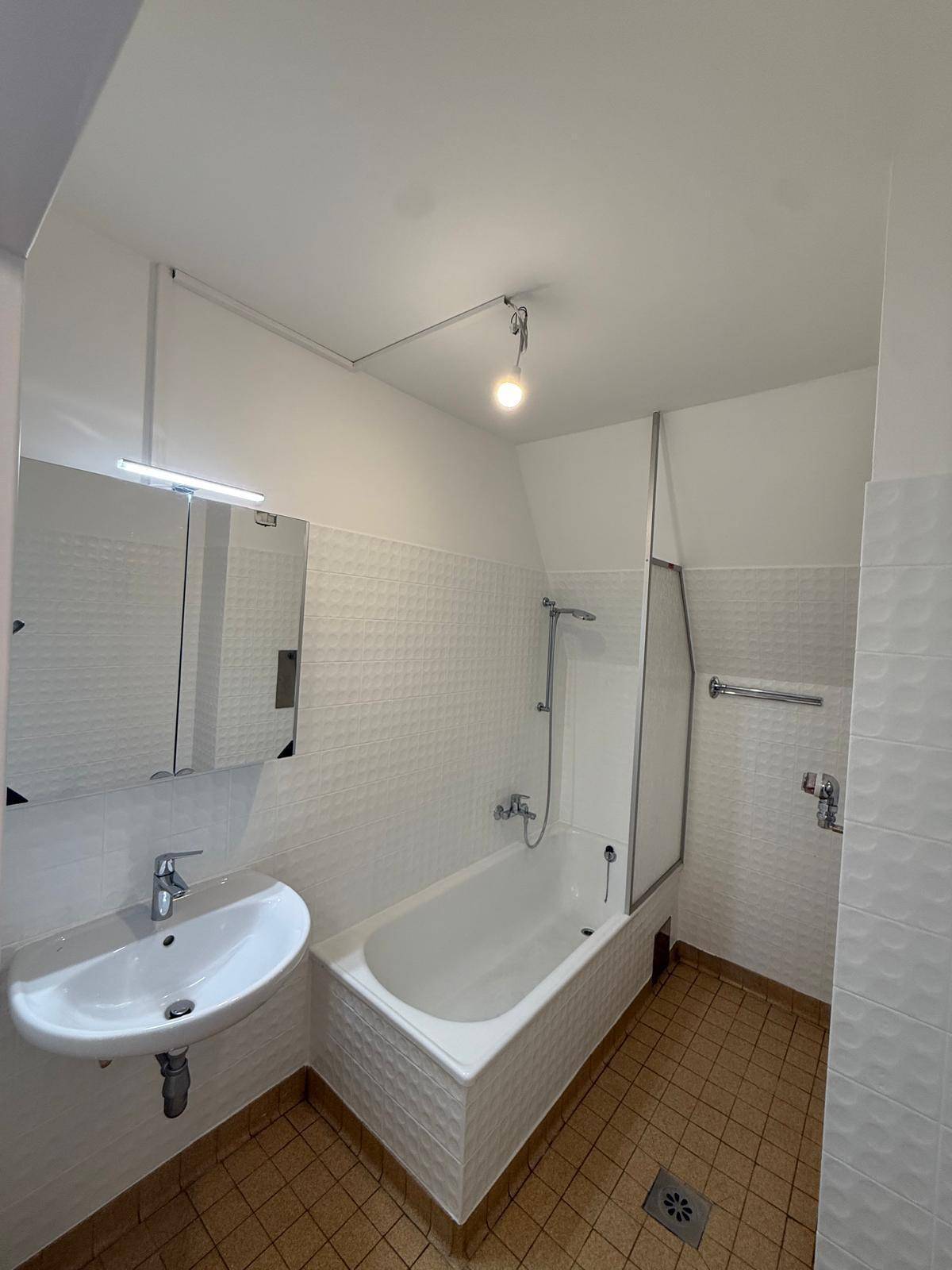 Badezimmer mit Badewanne inklusive Duschabtrennung, Waschbecken und Spiegelschrank, hell gefliest.