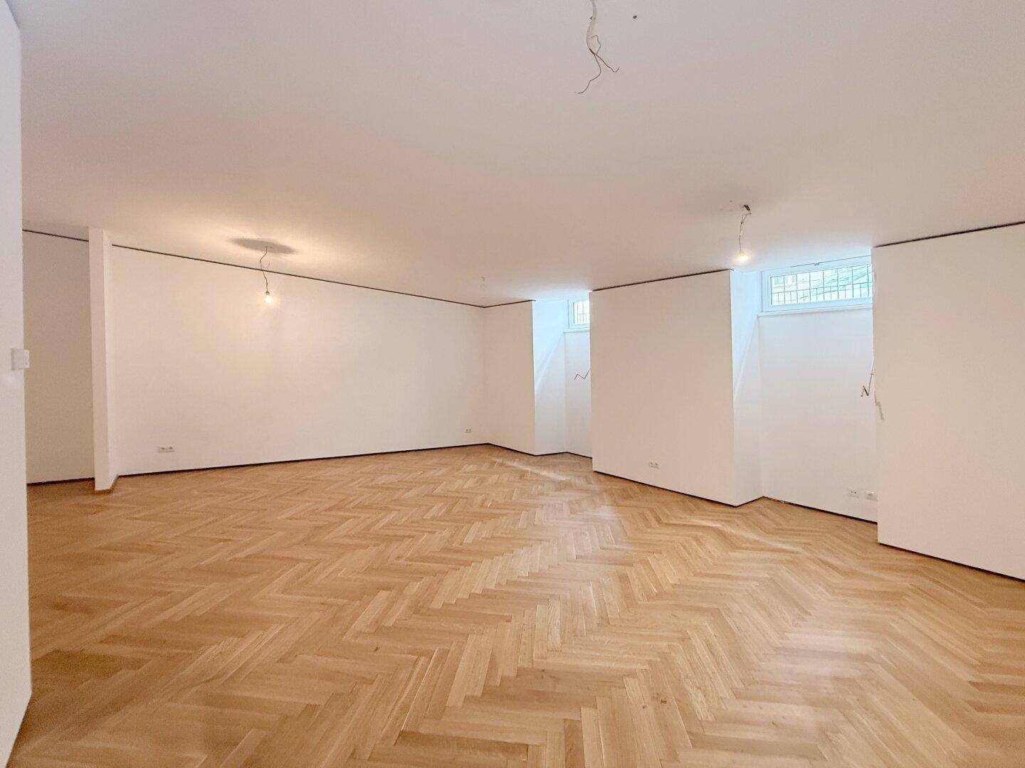 Leerer Raum mit Fischgrätparkett, weißen Wänden und hohen Fenstern nahe der Decke.