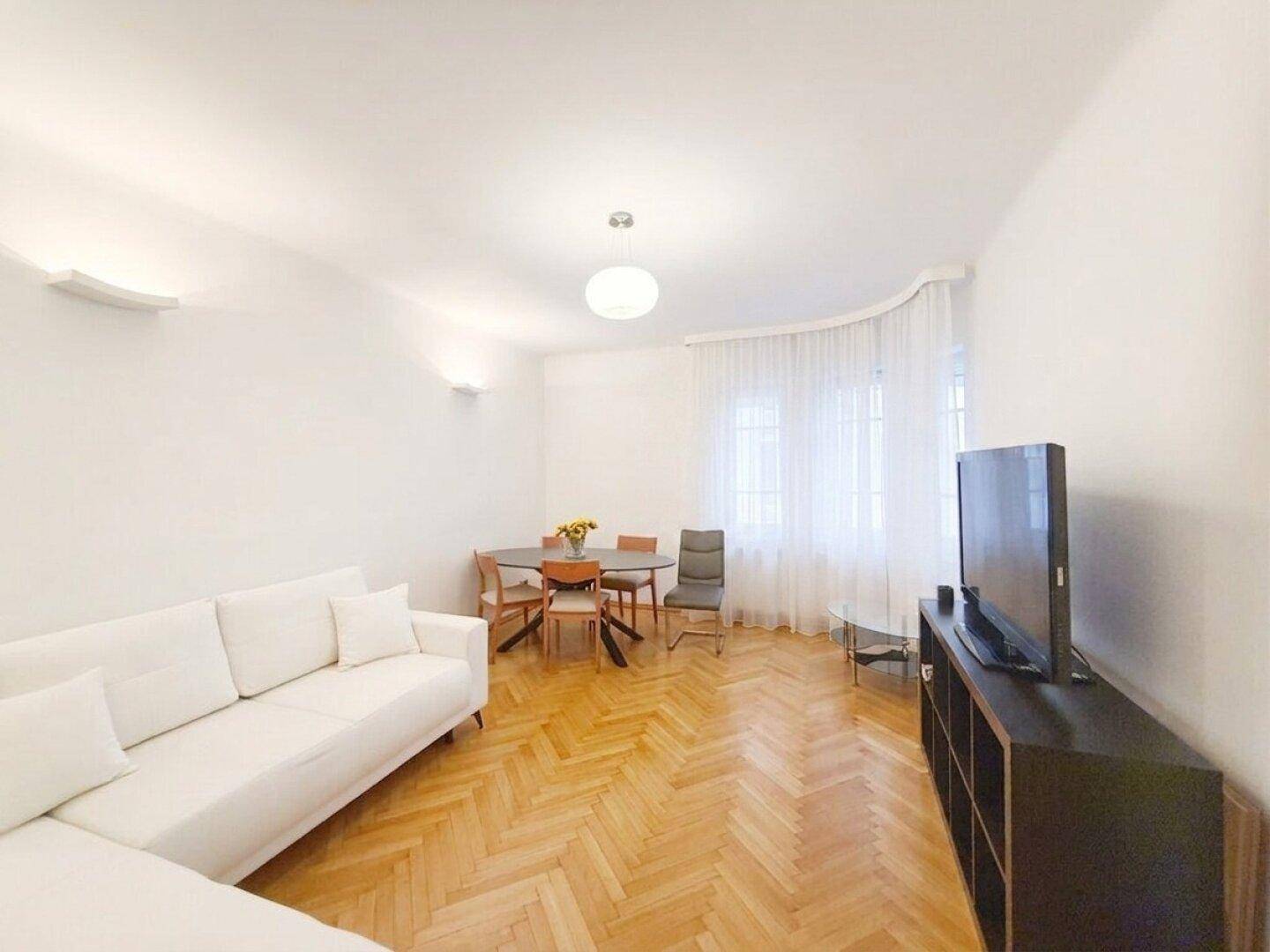 Helles Wohn- und Esszimmer mit Fischgrätparkett, weißem Sofa und einem Esstisch am Fenster.