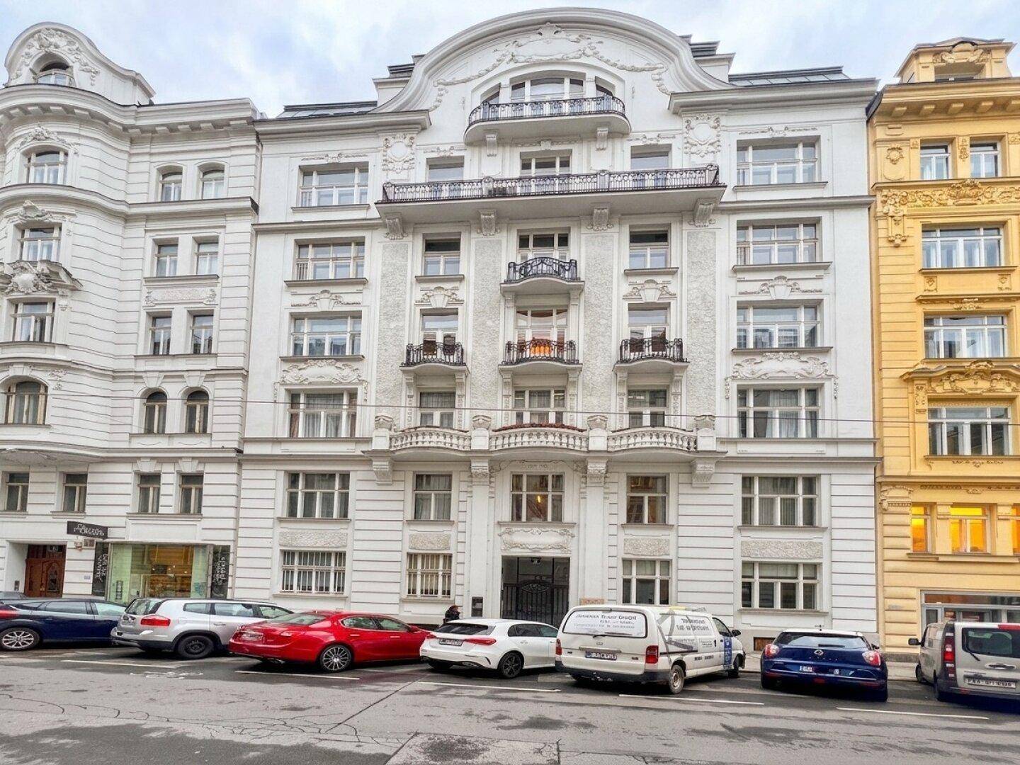 Elegante weiße Fassade eines Altbaus mit verzierten Balkonen und parkenden Autos davor.