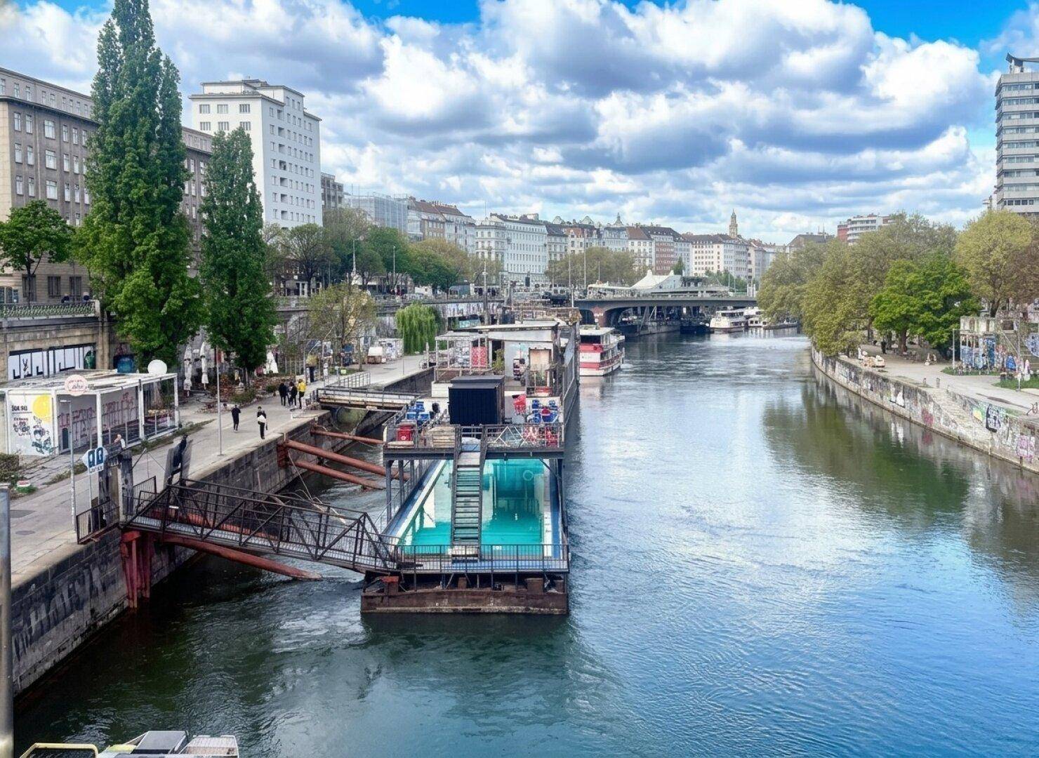 Flusslandschaft mit städtischer Bebauung und einem schwimmenden Pool unter bewölktem Himmel.
