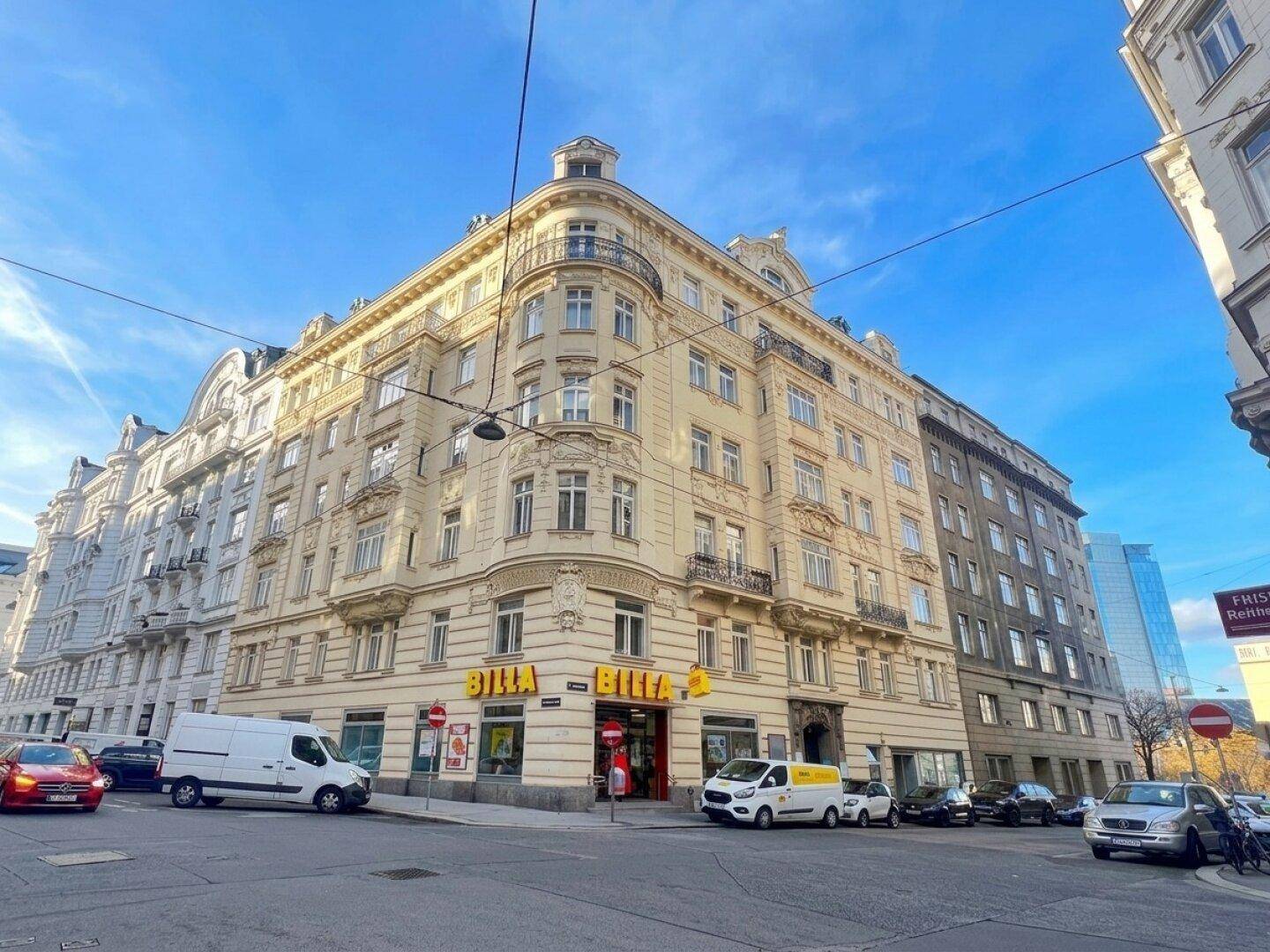 Eckgebäude mit Supermarkt im Erdgeschoss und belebter Straße unter blauem Himmel.