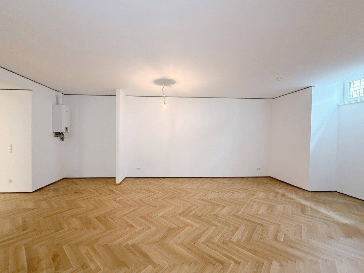 Leerer Raum mit Fischgrätparkett, weißen Wänden und einem an der Wand montierten Heizkessel.