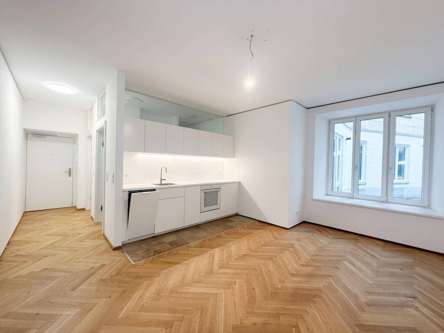 Offener Wohnbereich mit moderner weißer Küchenzeile, Fischgrätparkett und großen Fenstern.