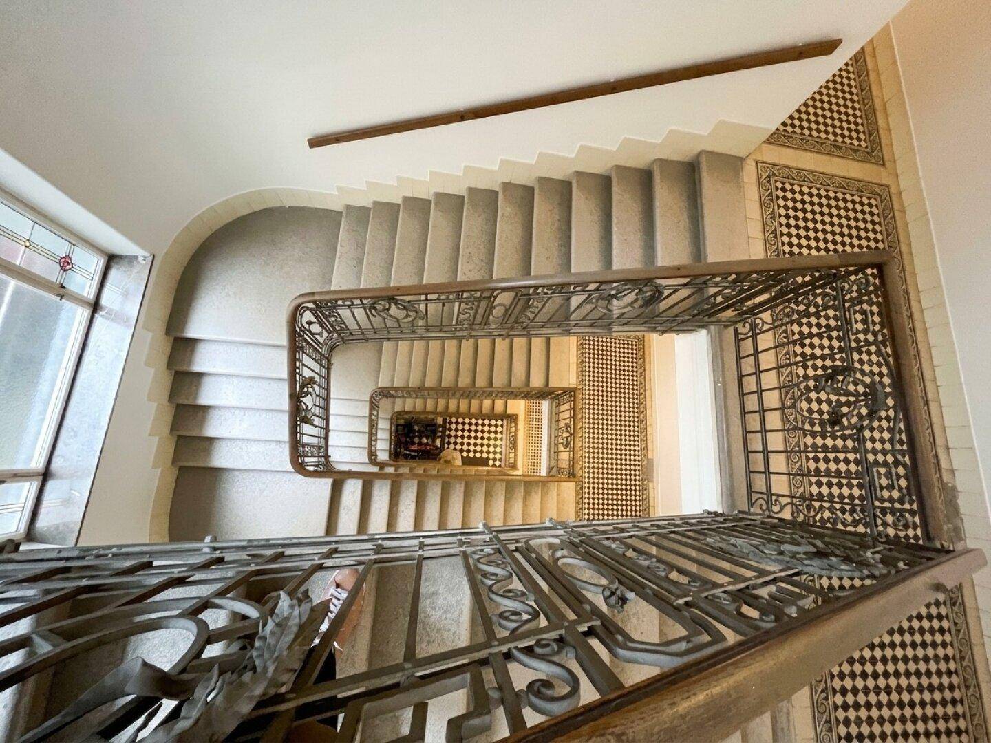 Blick von oben auf eine Wendeltreppe mit kunstvollen Geländern und gemusterten Bodenfliesen.