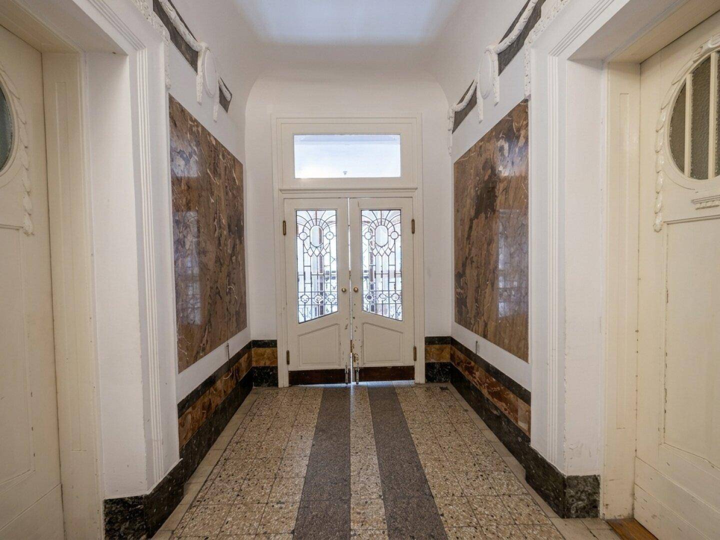Eingangsbereich eines Altbaus mit verzierten Türen, Marmorverkleidungen und gemustertem Fliesenboden.