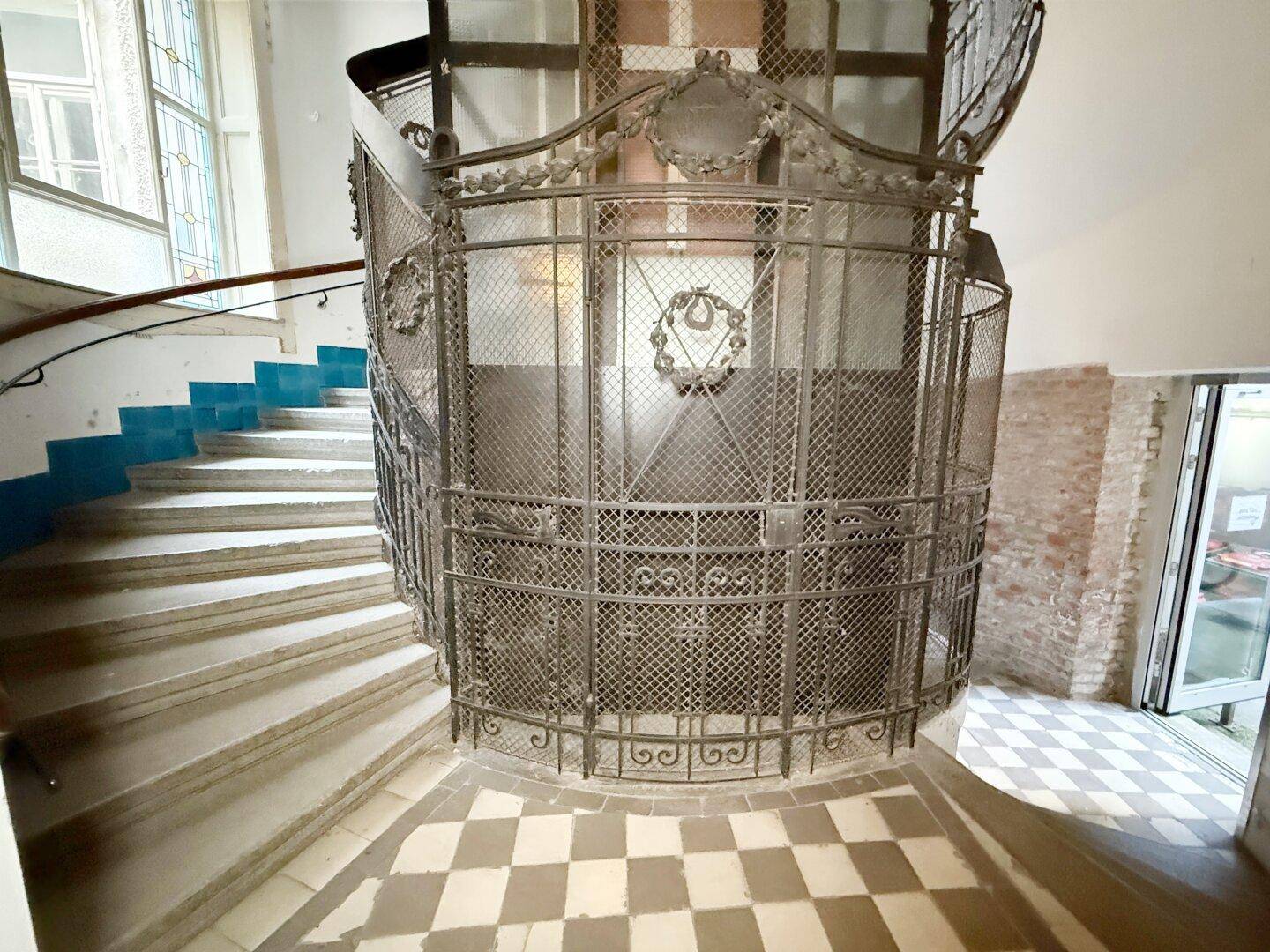 Historischer Gitteraufzug in einem Treppenhaus mit Wendeltreppe und kariertem Fliesenboden.