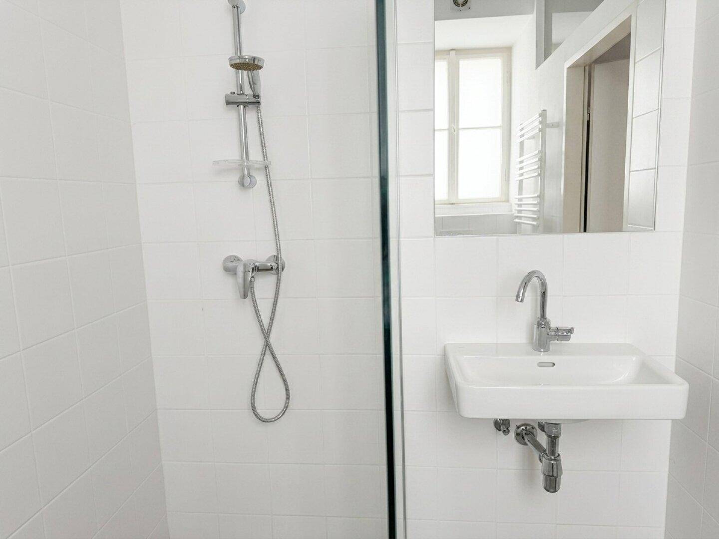 Modernes Badezimmer mit Duschkabine, weißen Fliesen und einem wandmontierten Waschbecken mit Spiegel.