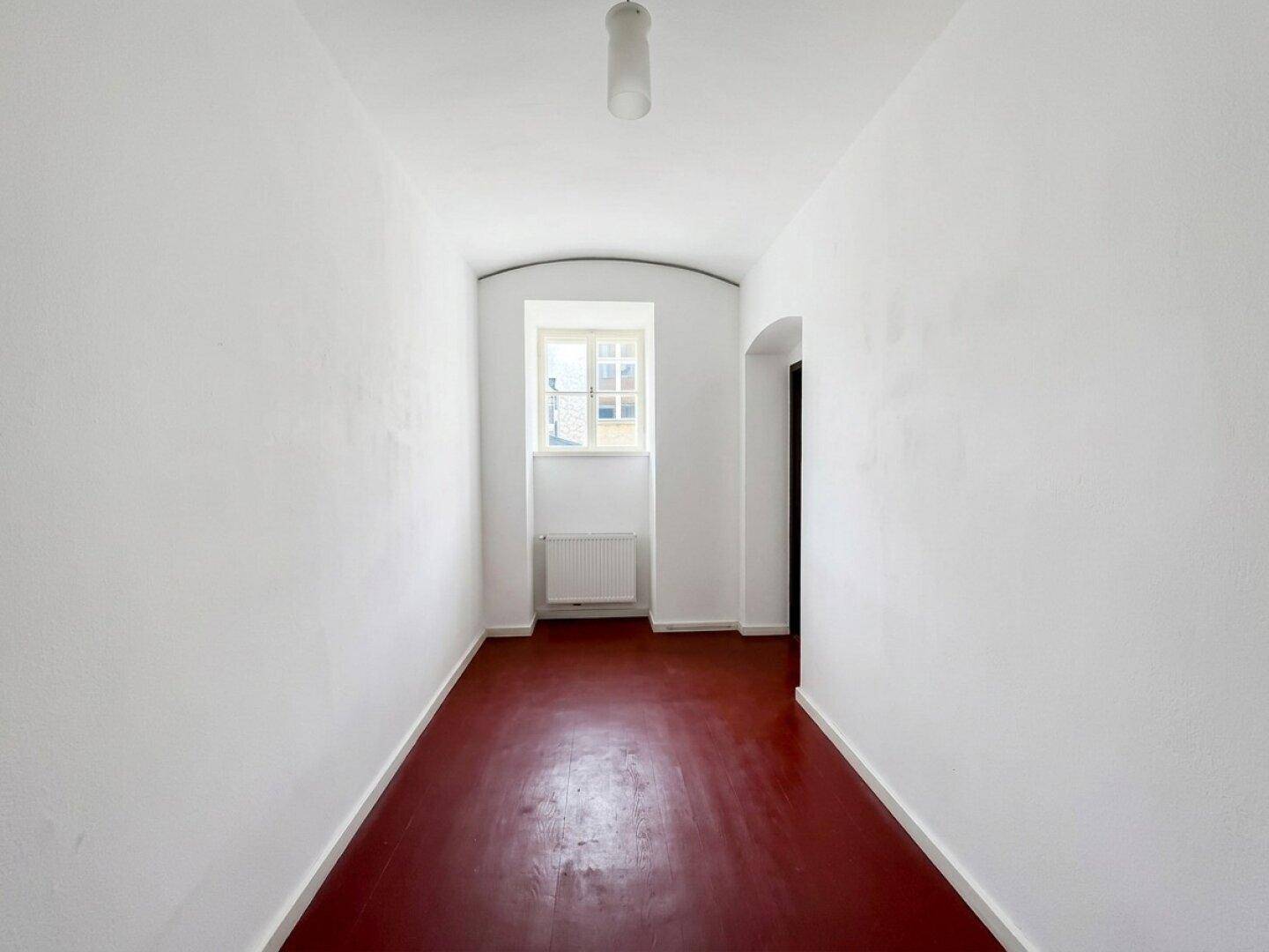 Langer, schmaler, leerer Raum mit dunkelrotem Boden und einem Fenster am Ende.