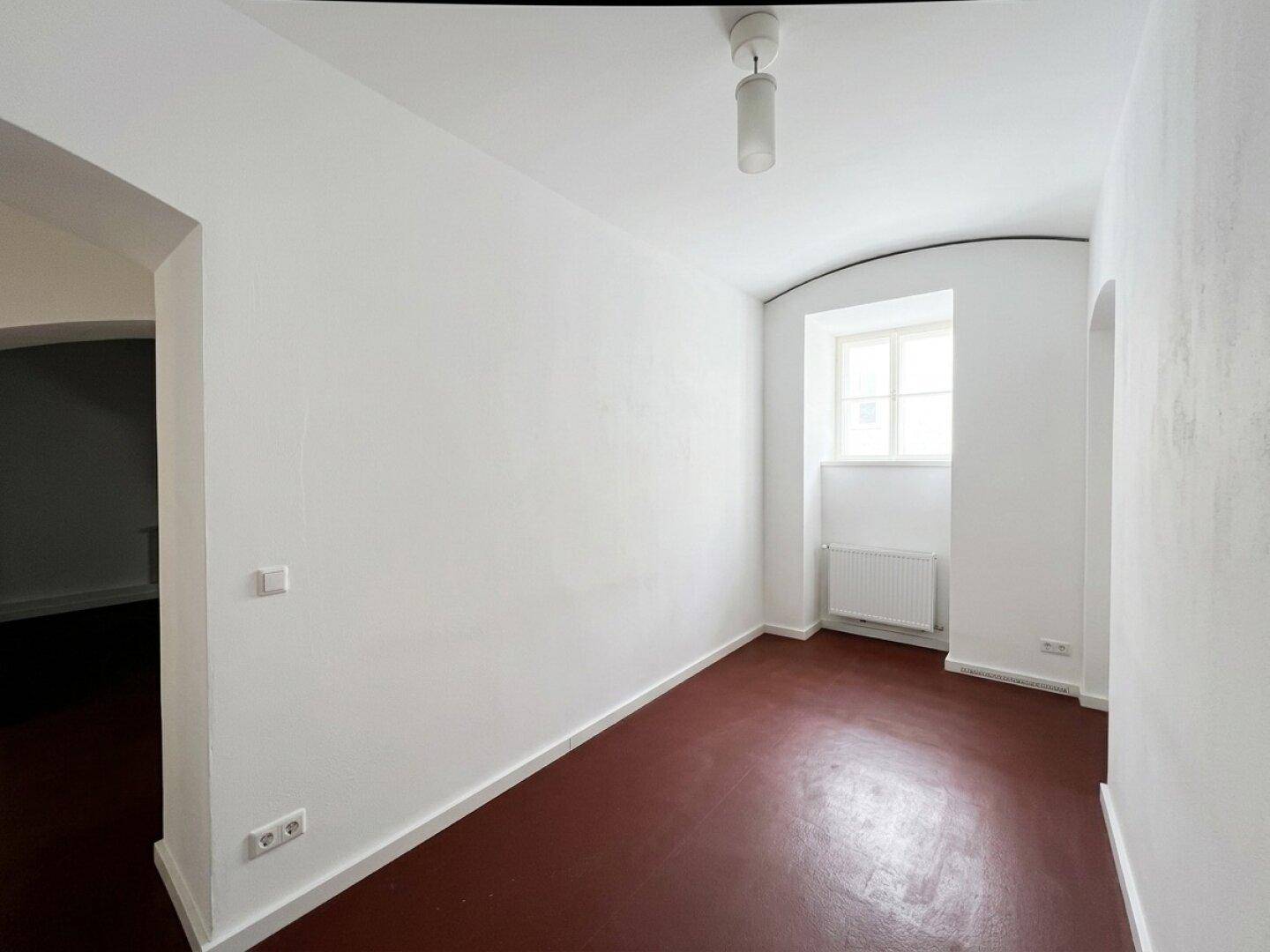 Leerer Raum mit dunkelrotem Boden, weißen Wänden und einem Fenster, das Tageslicht hereinlässt.